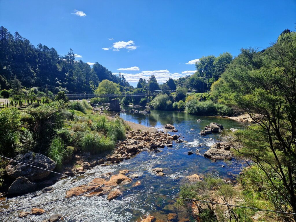 Image of Karangahake Gorge, a scenic location in New Zealand. A clear river winds through a rocky bed, flanked by lush greenery and dense native trees. A bridge stretches across the gorge, connecting the two sides. The sky is a brilliant blue with scattered clouds, enhancing the serene and picturesque atmosphere of the landscape