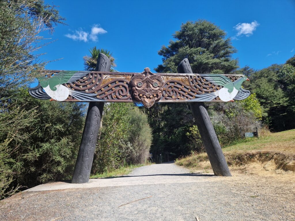 Image of a large wooden archway with intricate carvings at the entrance of a pathway surrounded by lush greenery. The carvings include a central mask-like figure with swirling patterns and two bird motifs on either side. The structure is supported by tall, dark wooden posts. The sky above is bright blue with scattered clouds, adding to the inviting and serene atmosphere of the scene.