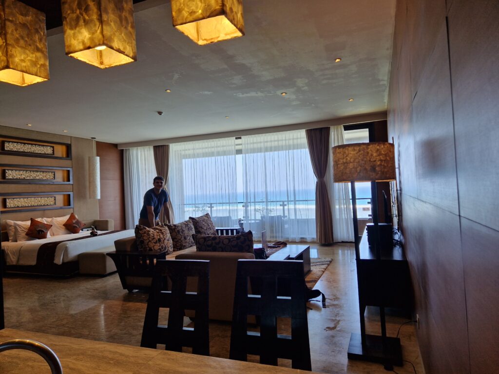 Image of a spacious and elegantly furnished living area in a hotel suite or luxury apartment. The room features a large bed adorned with decorative pillows, a plush sofa set with patterned cushions, and a wooden coffee table. A dining table with chairs is also present, accompanied by ambient lighting from pendant lamps and a floor lamp. The windows are dressed with sheer curtains, allowing natural light to filter in while offering a breathtaking view of the ocean in the background. The atmosphere is warm and inviting, creating a perfect setting for relaxation or vacation.