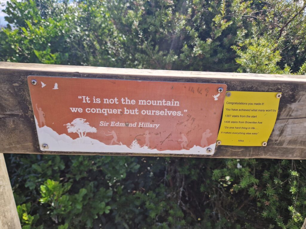 Image of a wooden fence with two attached motivational signs. The left sign is orange with white text and features a quote by Sir Edmund Hillary: 'It is not the mountain we conquer, but ourselves.' The right sign is yellow with black text, " Congratulations you made it! You have achieved what many wont try. 1387nstairs from the start, 1496 stairs from Brownlee Avenue. 'Do one hard thing in life... It makes everything else easy.' - Mike.