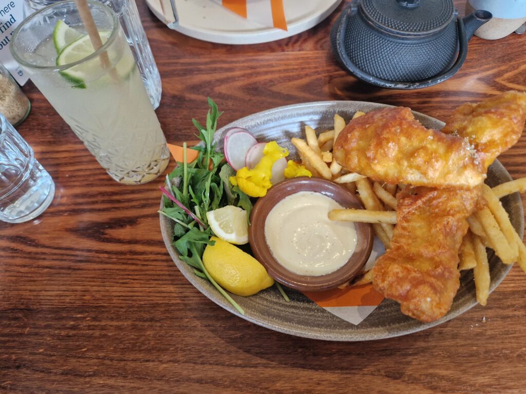 "Image of a classic fish and chips dish from Hayes Common. The plate features two golden-battered fish fillets alongside a generous serving of crispy French fries. Accompaniments include a small bowl of tartar sauce, a lemon wedge for added zest, and a fresh side salad of mixed greens, thinly sliced radishes, and vibrant pickled vegetables. The meal is presented on a rustic wooden table, paired with a refreshing glass of lemonade garnished with lime slices, creating an inviting and hearty dining experience.