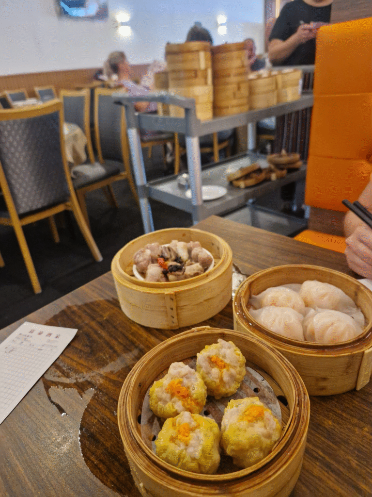 Image of a table at Regal Chinese Restaurant in Hamilton, featuring three bamboo steamers filled with dim sum dishes. One steamer contains siu mai dumplings, another holds har gow dumplings, and the third is filled with pork ribs cooked with black beans. A cart in the background displays additional bamboo steamers and plates, while other tables and chairs in the dining area are visible. A piece of paper with Chinese characters lies partially unfolded on the table, adding an authentic touch to the scene.
