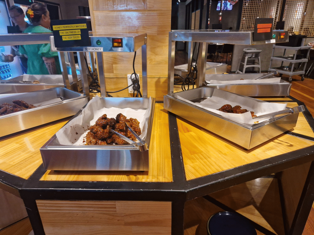 Image of a Korean fried chicken buffet in Tongkun. Three metal trays, each containing a different type or flavor of fried chicken, are placed on a wooden counter. The trays are equipped with heating lamps overhead to maintain the food's warmth. Tongs rest in the trays for easy self-service. The setup is clean and organized, with a cozy and casual atmosphere in the background, featuring people and kitchen equipment.