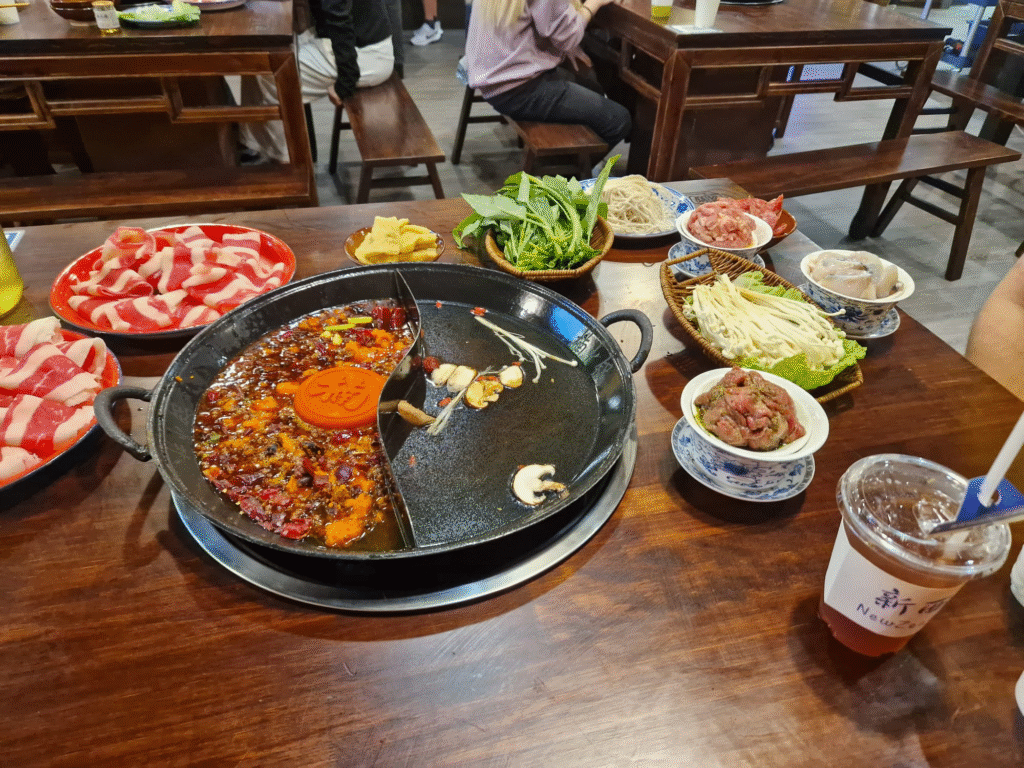 Image of a hot pot meal at a restaurant. At the center of the table is a divided hot pot with two types of broth: one is spicy beef tallow which has red colour with chili peppers, and the other is a clear broth with mushrooms. Surrounding the hot pot are plates of raw meat, bowls of fresh vegetables, mushrooms, tofu, and other ingredients ready to be cooked in the broths. A drink with a straw is also placed on the table. The ambiance of the restaurant includes wooden tables and chairs, creating a warm and traditional dining atmosphere