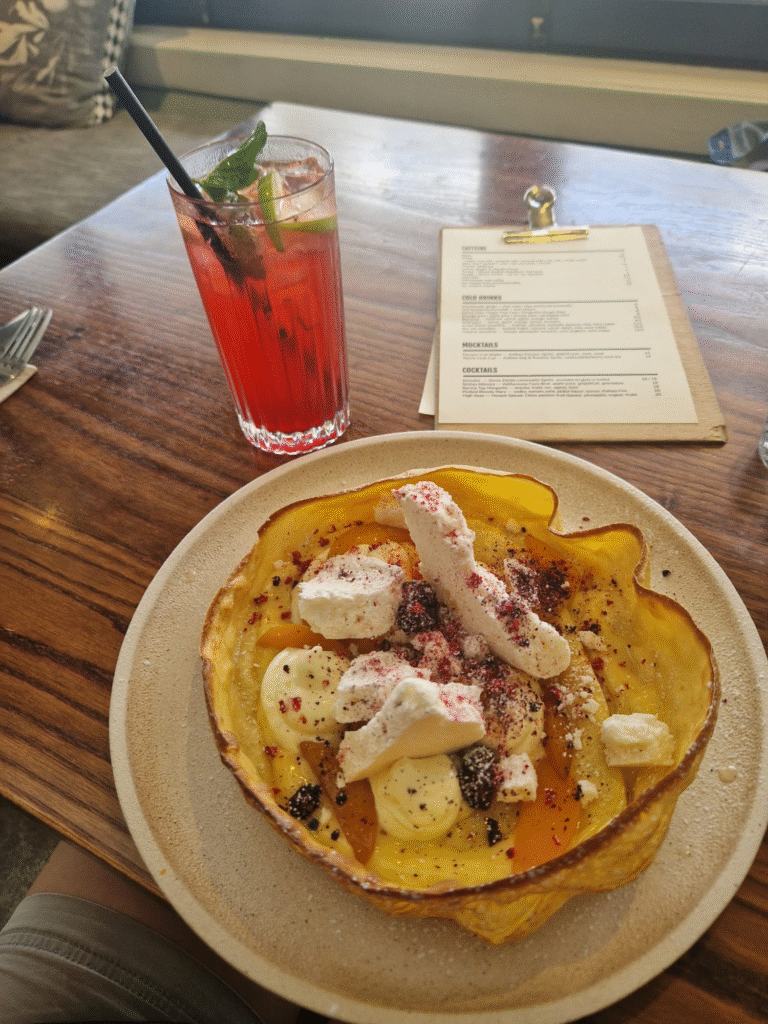 Image of a beautifully served Dutch baby pancake from Hayes Common. The dish is topped with meringue, fresh berries like strawberries and blueberries, and dusted with powdered sugar for a delicate finish. It is presented on a white plate set on a wooden table. Nearby, a tall glass of red iced drink garnished with mint leaves and served with a straw complements the meal. In the background, a clipboard menu adds a charming touch to the inviting setup