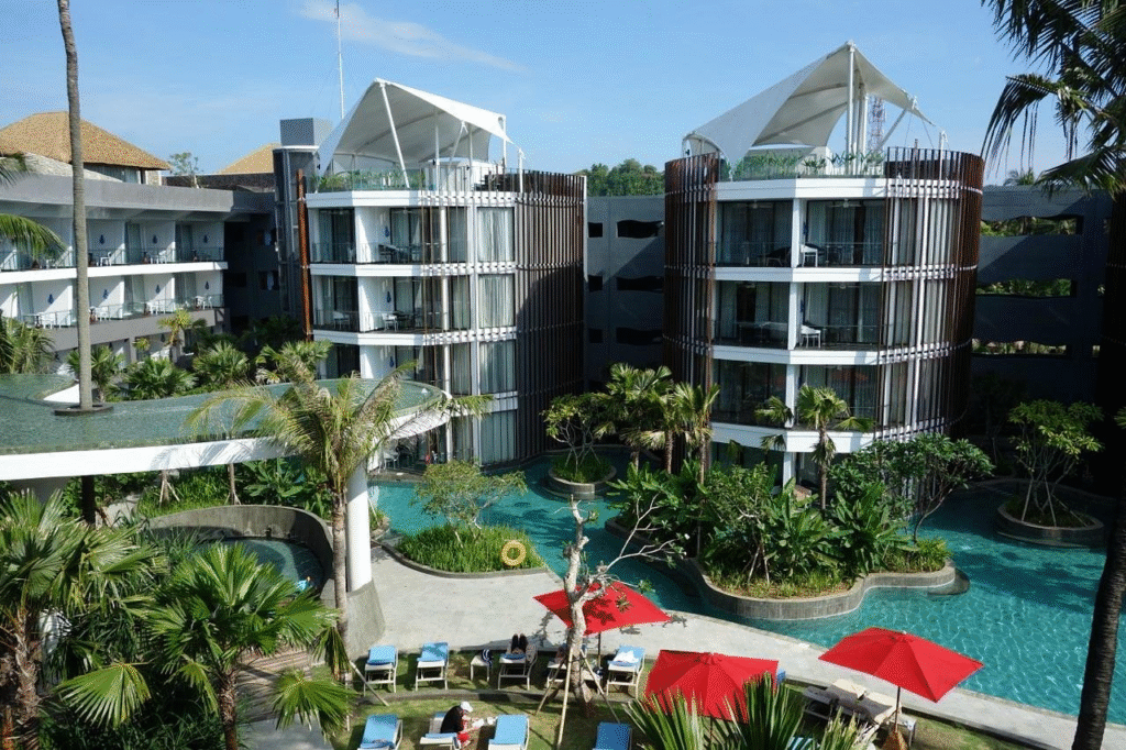 Image of a Le Meridien Jimbaran resort with two cylindrical buildings, each featuring multiple floors and balconies. The buildings are surrounded by lush tropical greenery, and a winding swimming pool flows between them. Red umbrellas and lounge chairs are positioned near the pool, creating a relaxing atmosphere. The resort's contemporary design blends clean architectural lines with natural elements, offering a luxurious and serene escape.