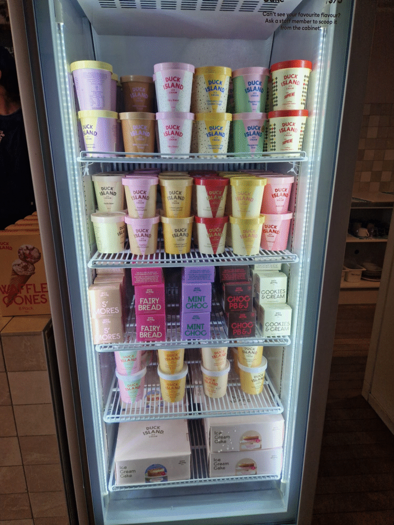 Image of a refrigerated display case featuring various Duck Island ice cream flavors. The containers are neatly arranged on shelves, showcasing a vibrant array of colors that represent flavors like S'mores, Fairy Bread, Mint Choc, Choc PBJ, and Cookies & Cream. A sign at the top reads: 'Can't see your favourite flavour? Ask a staff member to scoop it from the cabinet.' The display is bright and inviting, with clear labels on each container, offering a tempting selection for ice cream lovers.