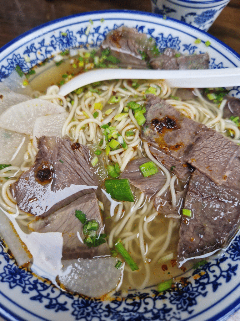 Image of a bowl of Lanzhou beef noodle soup featuring thinly sliced beef, Daikon slices, and springy noodles immersed in a clear, flavorful broth. The dish is garnished with finely chopped green onions for a pop of color, with a white ceramic soup spoon resting on the rim of the bowl. The bowl itself has an elegant blue and white floral pattern along its edge, adding a traditional touch to the presentation.