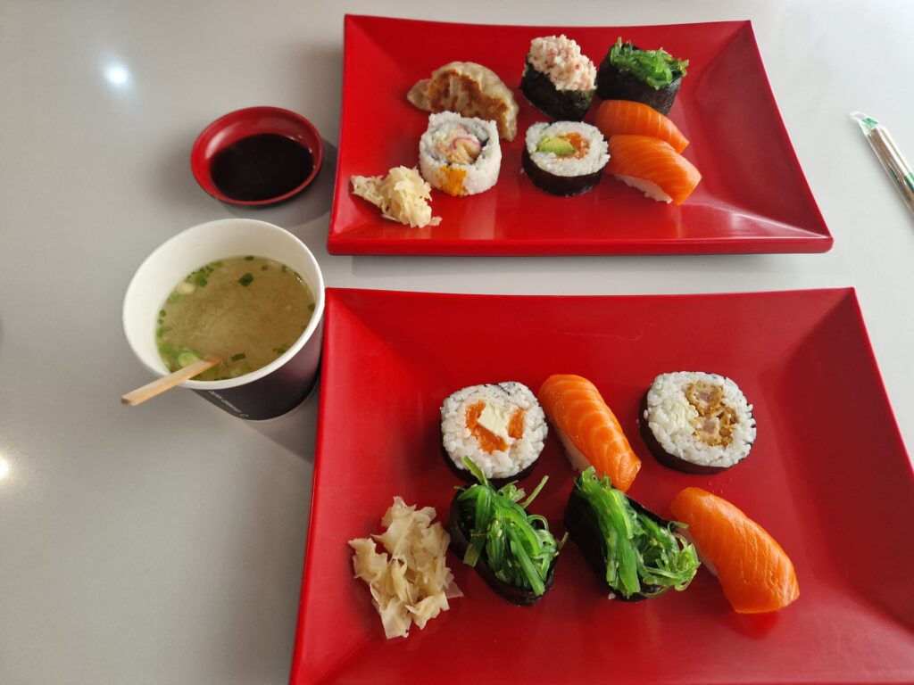 Image showcasing two red plates filled with a variety of sushi on a table. The upper plate features sushi nigiri with salmon, rolls with diverse fillings, and a small portion of ginger. The lower plate mirrors the contents of the top plate with similar sushi nigiri, rolls, and ginger. On the left, there's a bowl of miso soup with a wooden spoon resting beside it. Above the soup bowl is a small red dish containing soy sauce, adding to the inviting dining setup